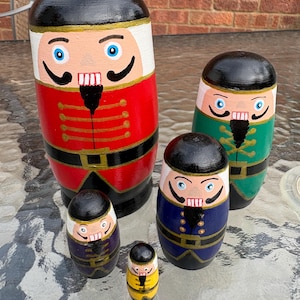 May include: A set of five wooden nesting dolls painted as nutcracker figures. The largest doll is red, followed by a green, a blue, a purple, and a yellow doll. Each figure features a black hat, blue eyes, a black moustache, and a white beard.