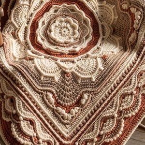 May include: A detailed, handmade crochet blanket in cream, brown, and rust colors. The blanket features a central floral design surrounded by intricate patterns and scalloped edges. The blanket is draped over a wooden chair.