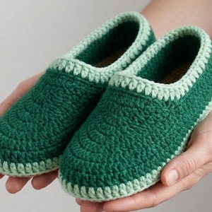 Può includere: Un paio di pantofole all'uncinetto verdi fatte a mano. Le pantofole hanno una punta arrotondata e un bordo verde chiaro attorno all'apertura e alla suola. Il punto all'uncinetto crea una superficie strutturata. Le pantofole sono tenute nelle mani di una persona.