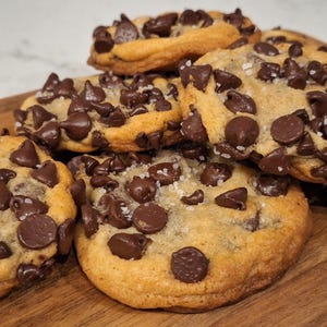 May include: A pile of freshly baked chocolate chip cookies on a wooden cutting board. The cookies are golden brown, generously studded with dark chocolate chips, and sprinkled with coarse salt.