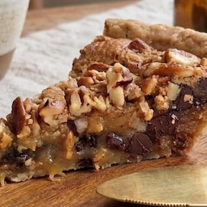 May include: A slice of pecan pie with a flaky crust, chocolate chips, and a caramel filling. The pie is topped with chopped pecans and sits on a wooden board. A gold pie server is next to the pie.