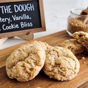 May include: Several light brown, textured cookies on a wooden board. A small chalkboard sign reads: JUST THE DOUGH - Buttery, Vanilla, Pure Cookie Bliss. A glass jar filled with brown sugar and a small wooden scoop sits behind the cookies.
