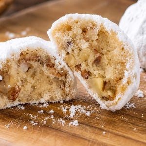 May include: A round cookie cut in half to reveal a light-colored, crumbly interior filled with visible chopped nut pieces. The exterior of the cookie is coated in a thick layer of white powdered sugar, with loose sugar scattered around the base.