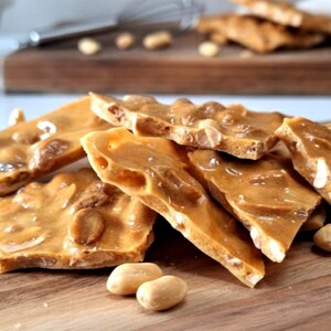 May include: Close-up of peanut brittle pieces on a wooden cutting board. The brittle has a golden-brown color with visible peanuts. Some loose peanuts are scattered around the brittle. The image suggests a homemade or artisanal treat.