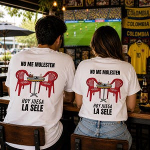 May include: Two people wearing white t-shirts with a graphic of a table and chairs, and the text "NO ME MOLESTEN HOY JUEGA LA SELE". The shirts are worn at a bar with a soccer game on TV.