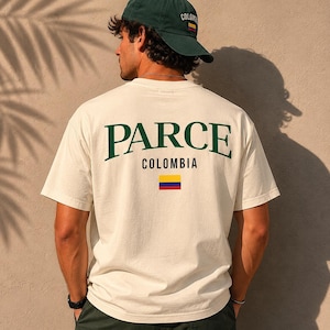 May include: Cream-colored t-shirt with the word "PARCE" in green arched lettering, "COLOMBIA" below, and the Colombian flag. A green baseball cap with "COLOMBIA" is worn. The shirt is short-sleeved.