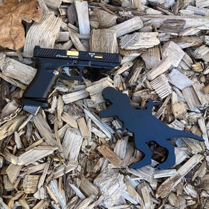 Peut inclure: Un pistolet noir avec des détails dorés et le texte "Combat Master" est présenté à côté d'un porte-clés en forme de dinosaure noir. Les objets reposent sur un lit de copeaux de bois, avec quelques feuilles.