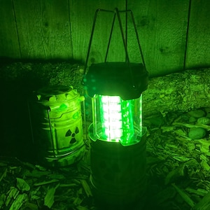 Puede incluir: Una linterna verde brillante con asa y base cilíndrica, junto a un contenedor verde a juego con un símbolo de radiación. La escena está iluminada con luz verde, lo que sugiere un tema postapocalíptico o de ciencia ficción.
