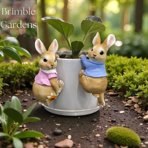 May include: A grey ceramic plant pot with a plant, decorated with two bunny figurines. One bunny wears a pink shirt, the other a blue shirt. The bunnies are positioned as if climbing the pot. The text "Brimble Gardens" is in the upper left corner.