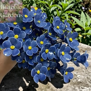 Puede incluir: Un ramo de flores de cerámica con pétalos azules y centros amarillos. Las flores están dispuestas sobre tallos negros. El texto "Brimble Gardens" está en la esquina superior izquierda.