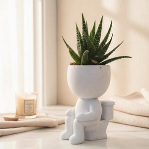 May include: A white ceramic planter in the shape of a person on a toilet, holding a green succulent plant. The planter has a speckled texture and is on a marble surface. A lit candle and folded towels are in the background.