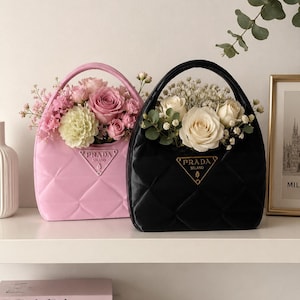 May include: Two handbag-shaped vases, one pink and one black, filled with flowers. The pink vase has a quilted design and a handle, with a Prada logo. The black vase has a similar design and handle, also with a Prada logo. The vases are on a white shelf.