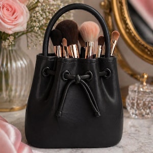May include: A black leather makeup brush holder with a handle, filled with various makeup brushes. The brushes have gold-coloured metal handles and different coloured bristles. The holder is on a marble surface, with a vase of pink roses in the background.