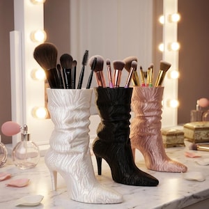 May include: Three high-heeled boot-shaped makeup brush holders in white, black, and pink, each filled with brushes. The boots have a textured design and are on a marble surface, with a lighted vanity mirror in the background.