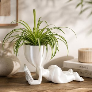 May include: A white, smooth-finished planter shaped like a human figure lying on its stomach whilst reading an open book. The head of the figure serves as the container for a green spider plant with long, thin, variegated leaves.