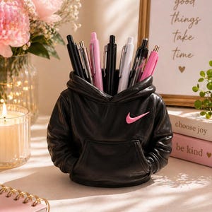 May include: A black hoodie-shaped pen holder with a pink Nike logo, filled with black, white, and pink pens. A framed print in the background reads "good things take time". The pen holder is a decorative desk accessory.