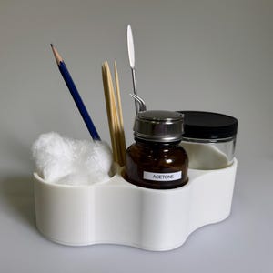 May include: A white multi-compartment organiser holding a blue pencil, wooden sticks, a metal spatula, a pair of tweezers, a ball of white cotton, and a brown glass bottle with a silver cap labelled ACETONE next to a jar with a black lid.