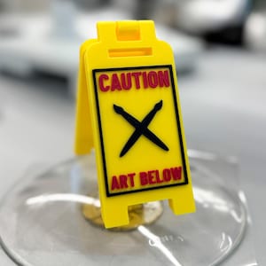 Op de afbeelding: Een geel plastic bord met het woord "CAUTION" in het rood en "ART BELOW" in het zwart. Het bord heeft een zwart "X"-symbool en is ontworpen om rechtop te staan, wat wijst op een mogelijk gevaar of interessegebied.