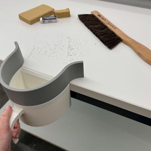 May include: A white and gray desk-mounted debris catcher with a handle is held by a hand. A wooden brush with dark bristles, a sanding block, and an eraser are on the white surface. The items suggest a workspace or craft area.