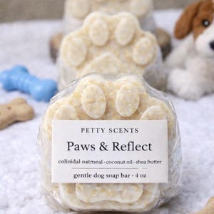 May include: A paw-shaped dog soap bar with the text "Paws & Reflect" on a white label. The soap is a light beige colour and is wrapped in clear plastic. The label also lists ingredients and the soap's weight, 113 g. Other dog-themed items are in the background.