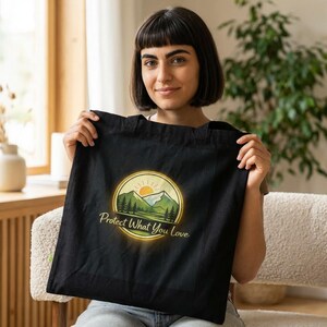 May include: Black tote bag with a circular graphic featuring a mountain scene, trees, and a rising sun. The text "Protect What You Love" is printed below the graphic. The bag is held up by a person.