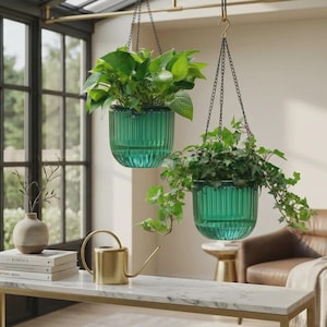 May include: Two hanging teal planters with lush green plants. The planters are suspended by black chains from a brass rod. A gold watering can and a stack of books sit on a marble-topped table.