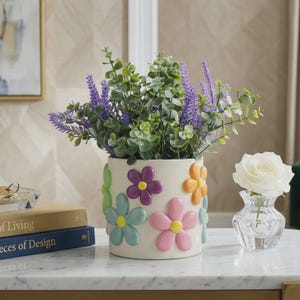 May include: A cream-colored ceramic flower vase with colorful 3D flower embellishments. The vase is filled with green and purple faux plants. A small glass vase with a white rose is to the right of the flower vase.