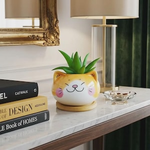May include: A ceramic cat-shaped planter with a green succulent plant. The planter is orange and white with a smiling cat face design. It sits on a wooden base, next to books and a lamp.