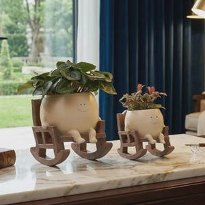 May include: Two whimsical, cream-colored planters shaped like smiling faces, each seated in a miniature wooden rocking chair. One planter holds leafy green plants, the other has red and green foliage. A charming decorative accent for any home.