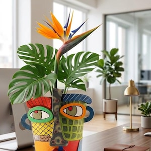 May include: A colorful, abstract face-shaped planter holding a Bird of Paradise flower and Monstera leaves. The planter features a yellow, red, green, and blue design with eye and lip details. The plant is on a wooden desk in a well-lit office.