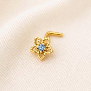 Op de afbeelding: Een gouden neusring met een bloemdesign met vijf bloemblaadjes. Het midden van de bloem heeft een kleine, ronde, lichtblauwe edelsteen. De neusring heeft een L-vormige pin.
