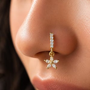 Puede incluir: Un aro de nariz dorado con un segmento articulado incrustado con una fila de pequeñas piedras redondas transparentes. Un dije colgante con forma de flor de cinco pétalos, engastado con piedras transparentes talla marquesa, cuelga del aro.