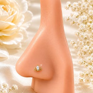 Può includere: Un piercing al naso dorato a forma di fiore di loto con una gemma centrale trasparente a taglio marquise. Il piercing è esposto su un manichino naso, con uno sfondo morbido e neutro e dettagli floreali.