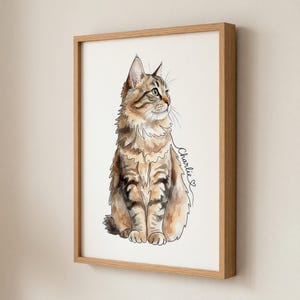 Op de afbeelding: Waterverfschilderij van een tabby kat genaamd Charlie, ingelijst in licht hout. De kat is afgebeeld in een zittende positie, met bruine en bruine vacht, groene ogen en een pluizige staart. De kat kijkt naar rechts.