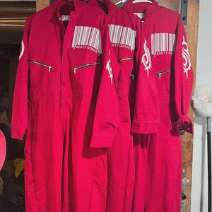 Puede incluir: Tres monos rojos colgados en una percha. Cada mono tiene una cremallera, un código de barras y un gráfico blanco en la manga. Los monos están hechos de un material resistente y están diseñados para el trabajo o el disfraz.