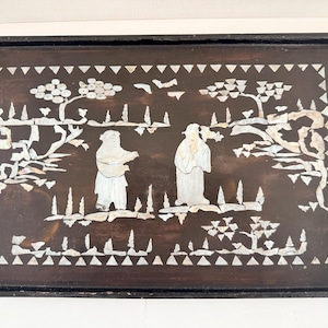 May include: A rectangular dark brown wooden tray featuring an inlaid mother-of-pearl design. The center depicts two figures standing among stylized trees and foliage, surrounded by a decorative border of small white triangular shapes.