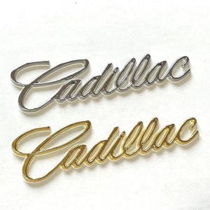 Puede incluir: Dos emblemas de Cadillac, uno plateado y otro dorado, se muestran sobre un fondo blanco. La escritura cursiva del logotipo de Cadillac es el punto focal, mostrando el diseño icónico de la marca. Los emblemas son probablemente para uso automotriz.