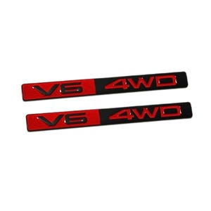 May include: Two rectangular automotive emblems featuring a split design. The left side is red with black V6 lettering, and the right side is black with red 4WD lettering. These badges have a raised, three-dimensional appearance with clean edges.