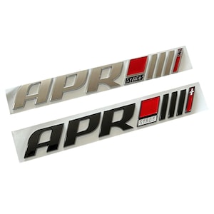 Op de afbeelding: Twee auto-embleemstickers. Eén is zilver met rode accenten, de andere is zwart met rode accenten. Beide hebben de letters "APR" in een vetgedrukte letter, met "STAGE" en verticale lijnen.