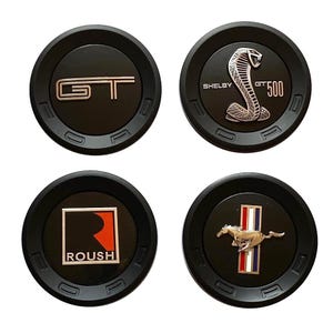 Puede incluir: Cuatro emblemas de coche negros con detalles plateados y rojos. Los emblemas presentan el logotipo GT, una cobra Shelby, el logotipo Roush y un caballo corriendo con rayas verticales. Cada emblema tiene un borde negro.