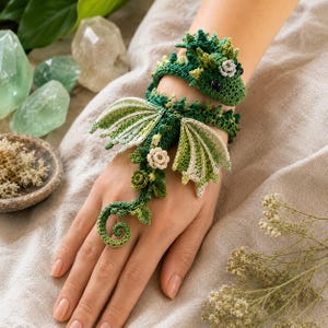 Può includere: Un braccialetto a forma di drago lavorato all'uncinetto in verde, con una testa dettagliata, occhi neri, squame testurizzate e due ali verde chiaro. Il corpo si estende sulla mano, decorato con piccoli fiori e foglie all'uncinetto color crema.