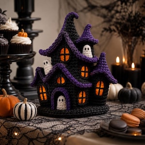May include: A crocheted haunted house decoration in black and purple yarn, featuring multiple pointed turrets, orange-lit windows, and three small white ghost figures. The structure has a textured, handmade appearance with detailed stitching.