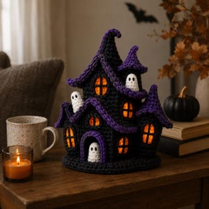 May include: A crocheted haunted house decoration in black and purple yarn with multiple pointed roofs. The structure features several windows glowing with orange light and three small white crocheted ghosts positioned in the doorway and on the roof.