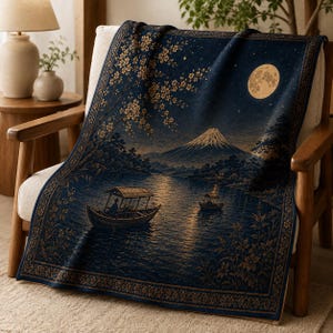 Op de afbeelding: Een donkerblauwe geweven plaid met een gedetailleerd landschap met een volle maan, de berg Fuji, kersenbloesemtakken en twee kleine bootjes op een meer. De plaid heeft een decoratief bloemenpatroon langs alle vier de randen.