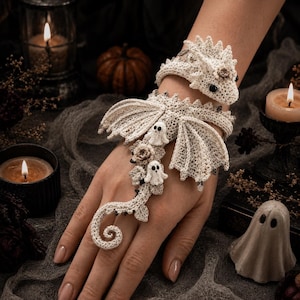 Könnte beinhalten: Ein beigefarbenes Häkel-Handornament mit einem Drachenkopf am Handgelenk, Flügeln über dem Handrücken und einem langen Schwanz, der sich um einen Finger wickelt. Das Stück ist mit kleinen gehäkelten Geisterfiguren und Blumenakzenten verziert.