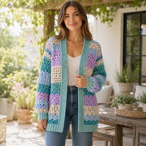May include: A colorful, open-front crochet cardigan in shades of blue, teal, purple, and cream. The long-sleeved sweater features a patchwork design with a turquoise border. The model is wearing the cardigan with a white top and blue jeans.