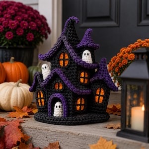 May include: A crocheted haunted house decoration featuring a black structure with purple roof accents, multiple orange-lit windows, and three small white ghost figures positioned at the entrance and on the upper levels of the building.