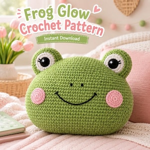 Puede incluir: Un cojín verde tejido a ganchillo con forma de rana, con mejillas rosas y grandes ojos negros y blancos. El texto "Frog Glow Crochet Pattern" y "Instant Download" se muestra en la imagen. El cojín está sobre una superficie rosa.