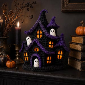 May include: A crocheted haunted house decoration in black and purple yarn, featuring multiple pointed roofs, arched windows with glowing orange light, and three small white ghost figures positioned on the exterior walls and at the entrance.