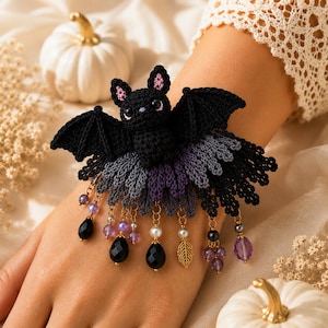 May include: A crochet bracelet featuring a black bat with pink inner ears and large eyes. The bat is attached to a layered, fan-like base in shades of black, gray, and purple. Dangling from the base are gold-tone chains with black and purple beads.
